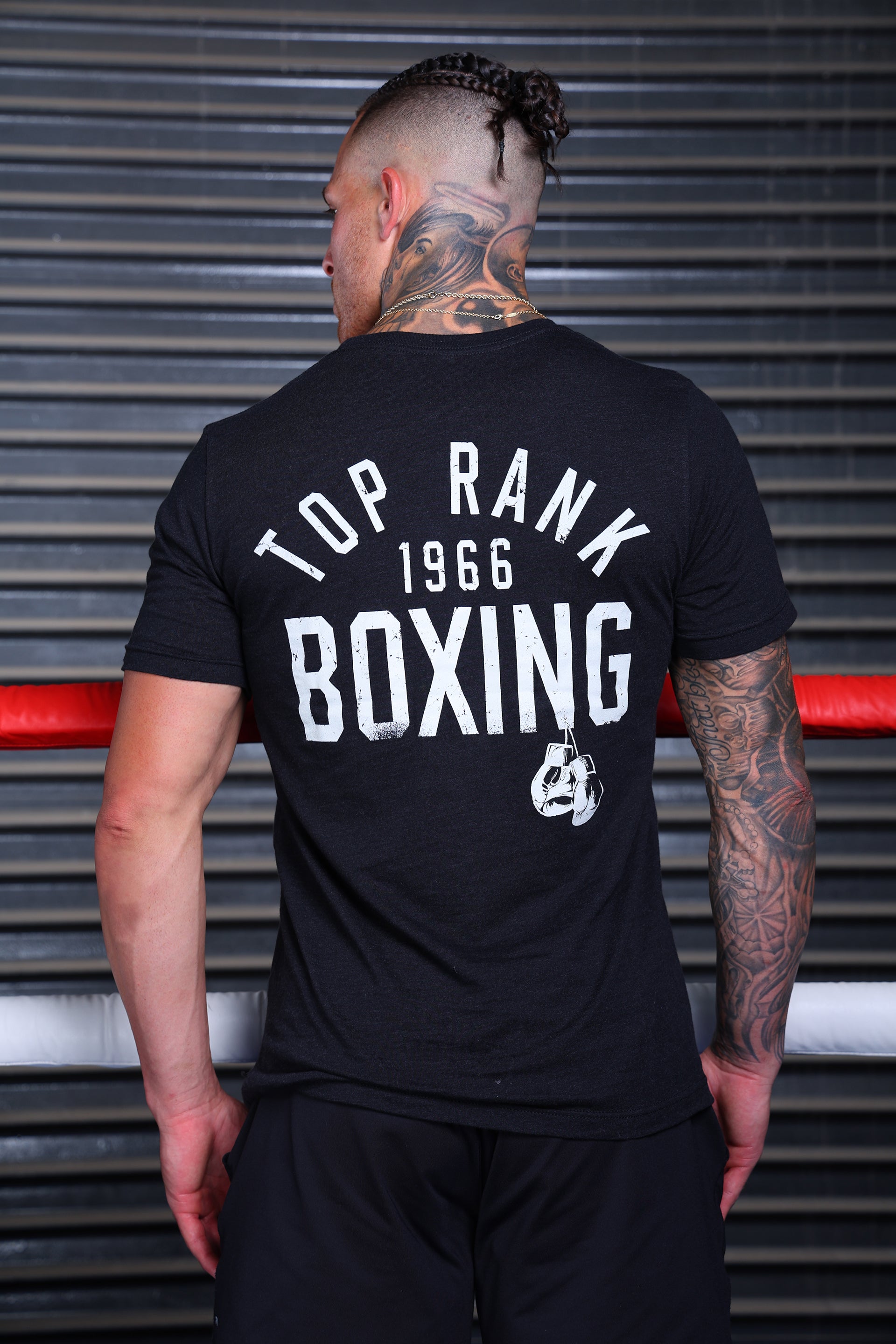 Back view of a man wearing a black t-shirt with "Top Rank 1966 Boxing" printed in white, standing in front of a boxing ring.